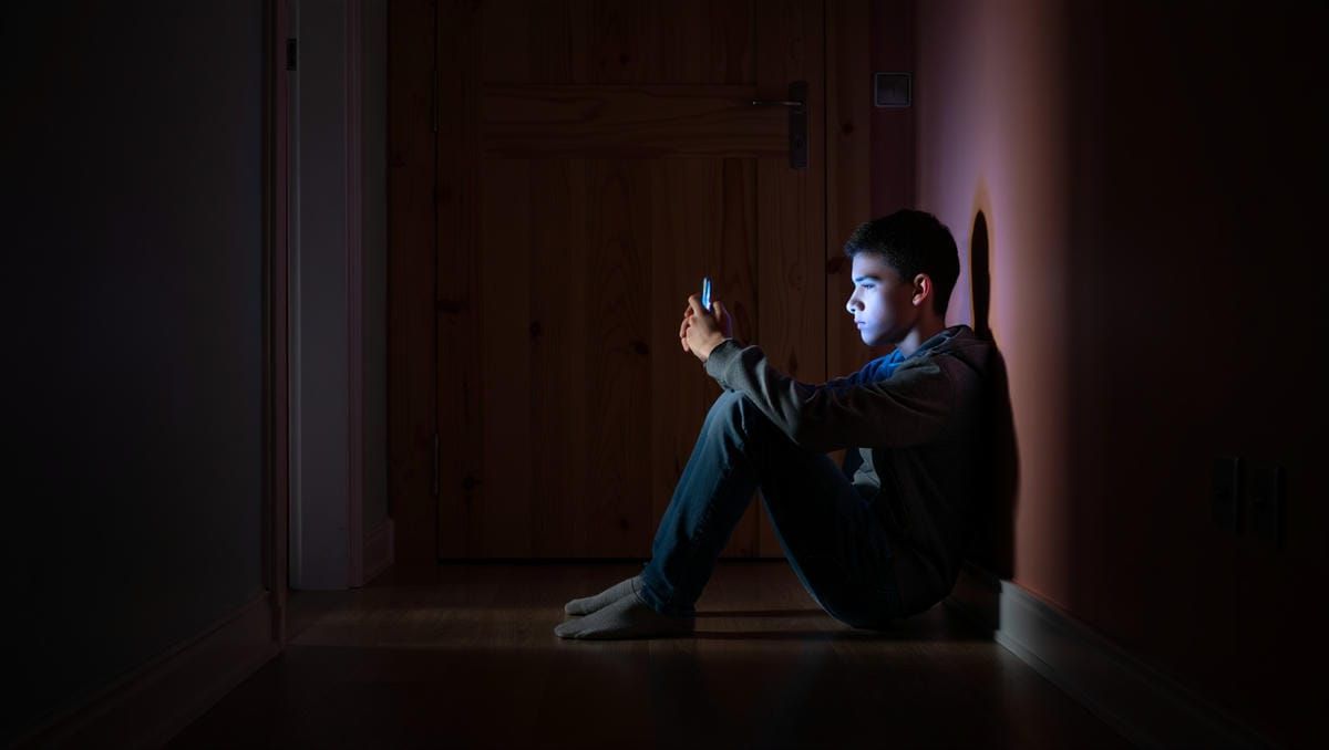 Подросток сидит на полу у закрытой двери в комнату, подсвеченной синим светом смартфона.