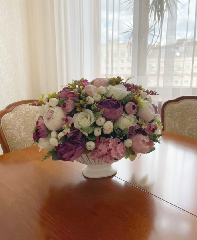 Композиция удлинённая из искусственных цветов в вазе лодочке на деревянном столе  на фоне окна