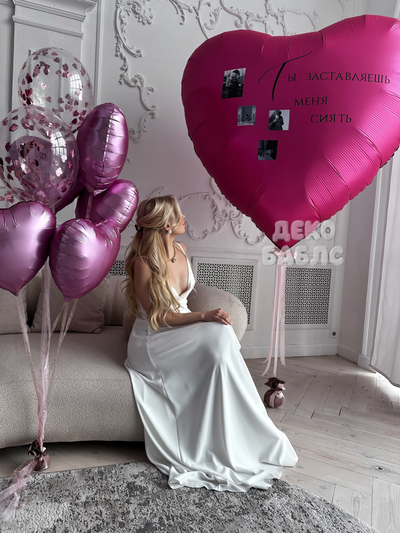 Девушка в белом платье рядом с гигантским малиновым сердцем, украшенным личными фото, и фонтаном из