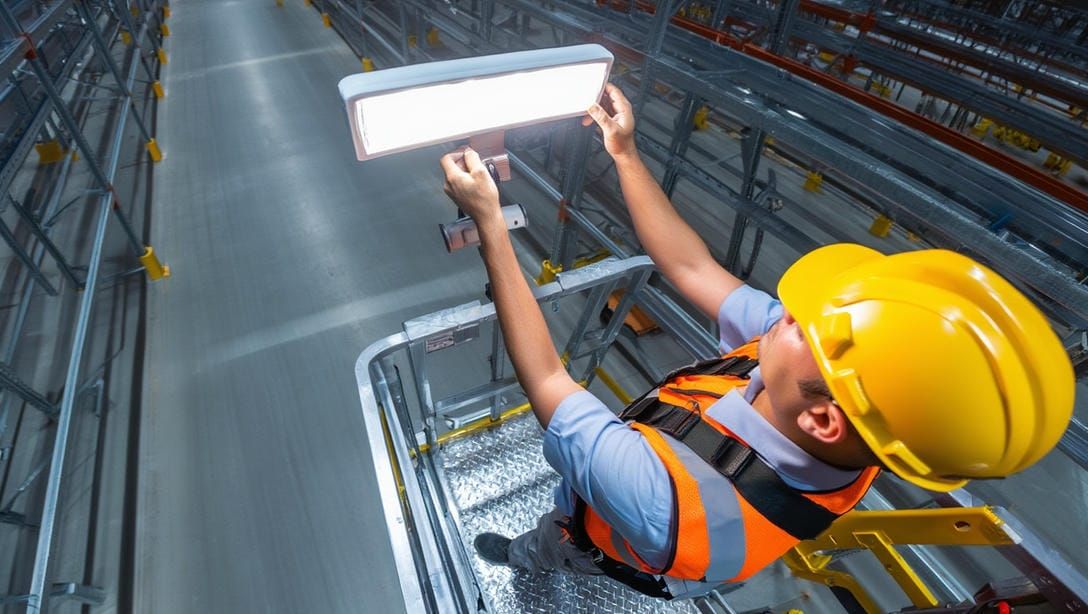 Техник устанавливает промышленный LED‑светильник на высоту стеллажей с подъёмником.