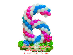 Плетеная цифра 6 из розово/бело/голубых шаров, расположенная на цветочной полянке зеленого цвета